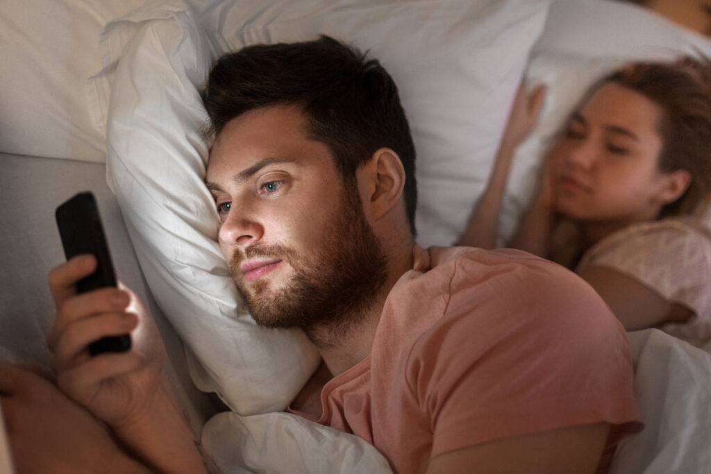 Man laying in bed looking on his phone with a woman behind him sleeping.