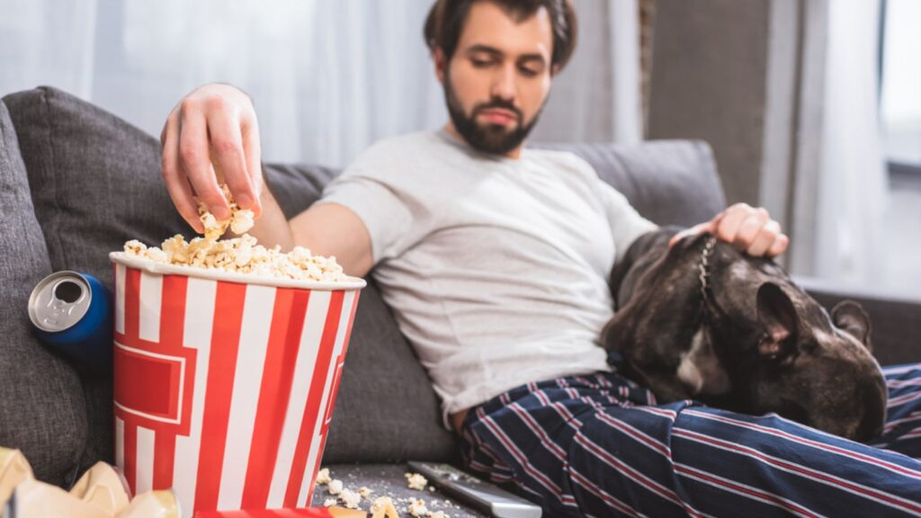 A man Sitting on a dirty Sofa, Eating Popcorns and His Pet sleeping on his lap