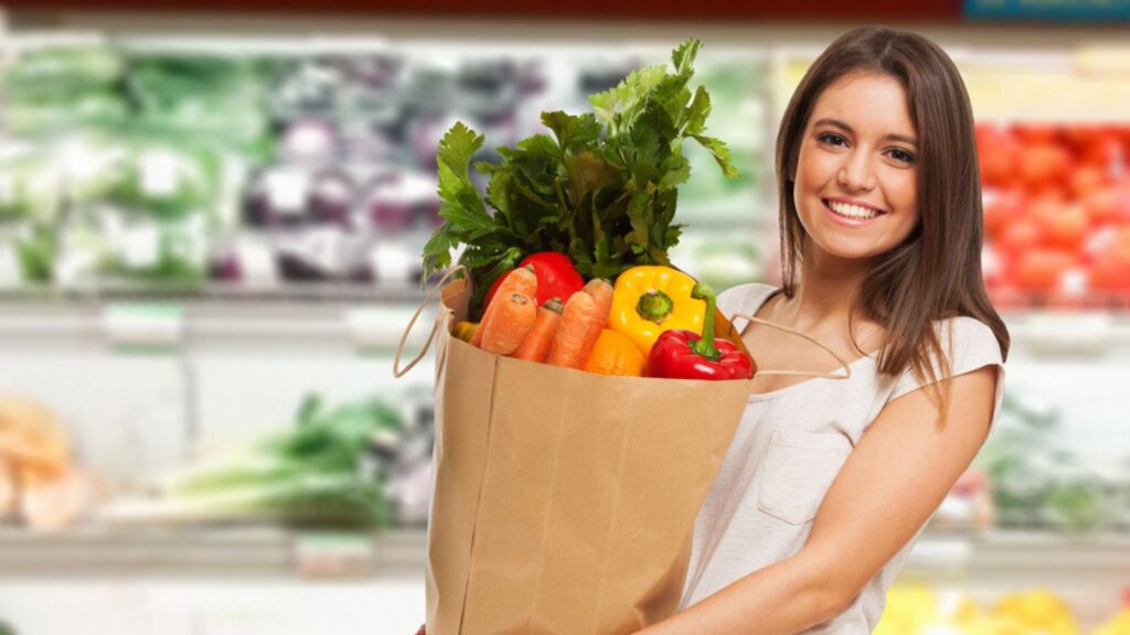 A woman smiling and standing in a grocery store, Holding a Waste free bag filled with Vegetables