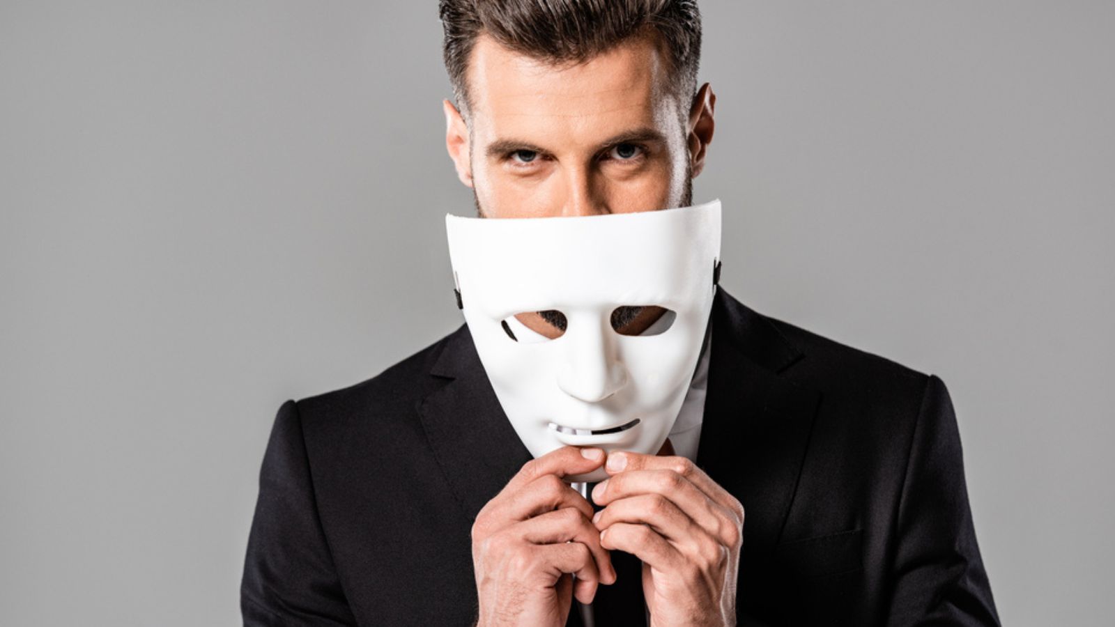Sly handsome businessman in black suit taking off white mask isolated
