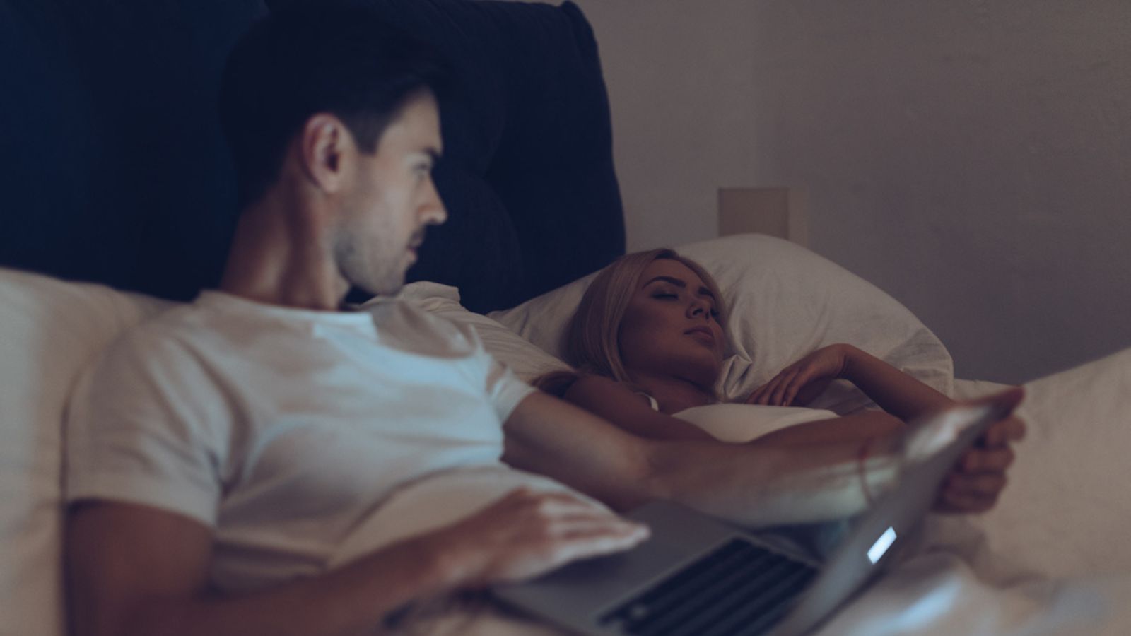 Suspicious young man using laptop and looking at girlfriend sleeping