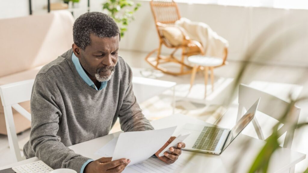 older man looking at his bills at his desk by his computer
