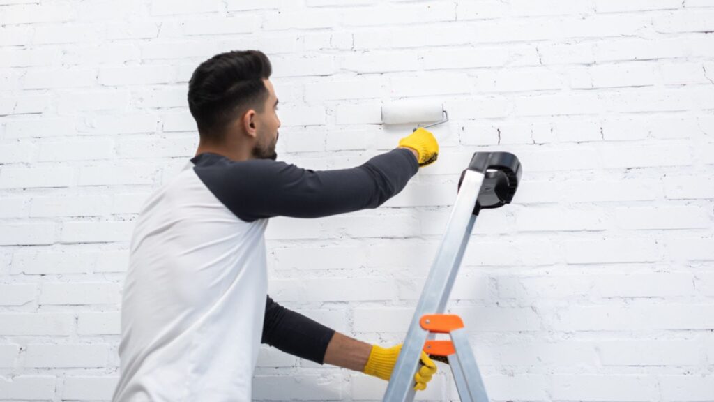 Young man painting wall with roller while standing on ladder