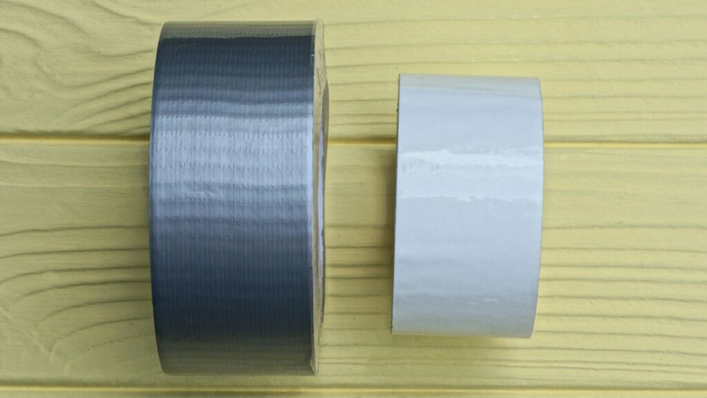 A roll of gray reinforced tape and a roll of white tape lie on a yellow wooden table
