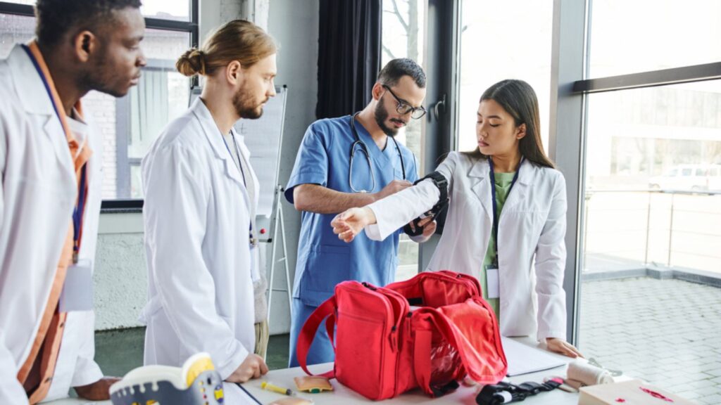Healthcare worker in eyeglasses and uniform applying compressive tourniquet on arm of asian woman near multiethnic students, first aid kit and medical equipment, bleeding prevention concept