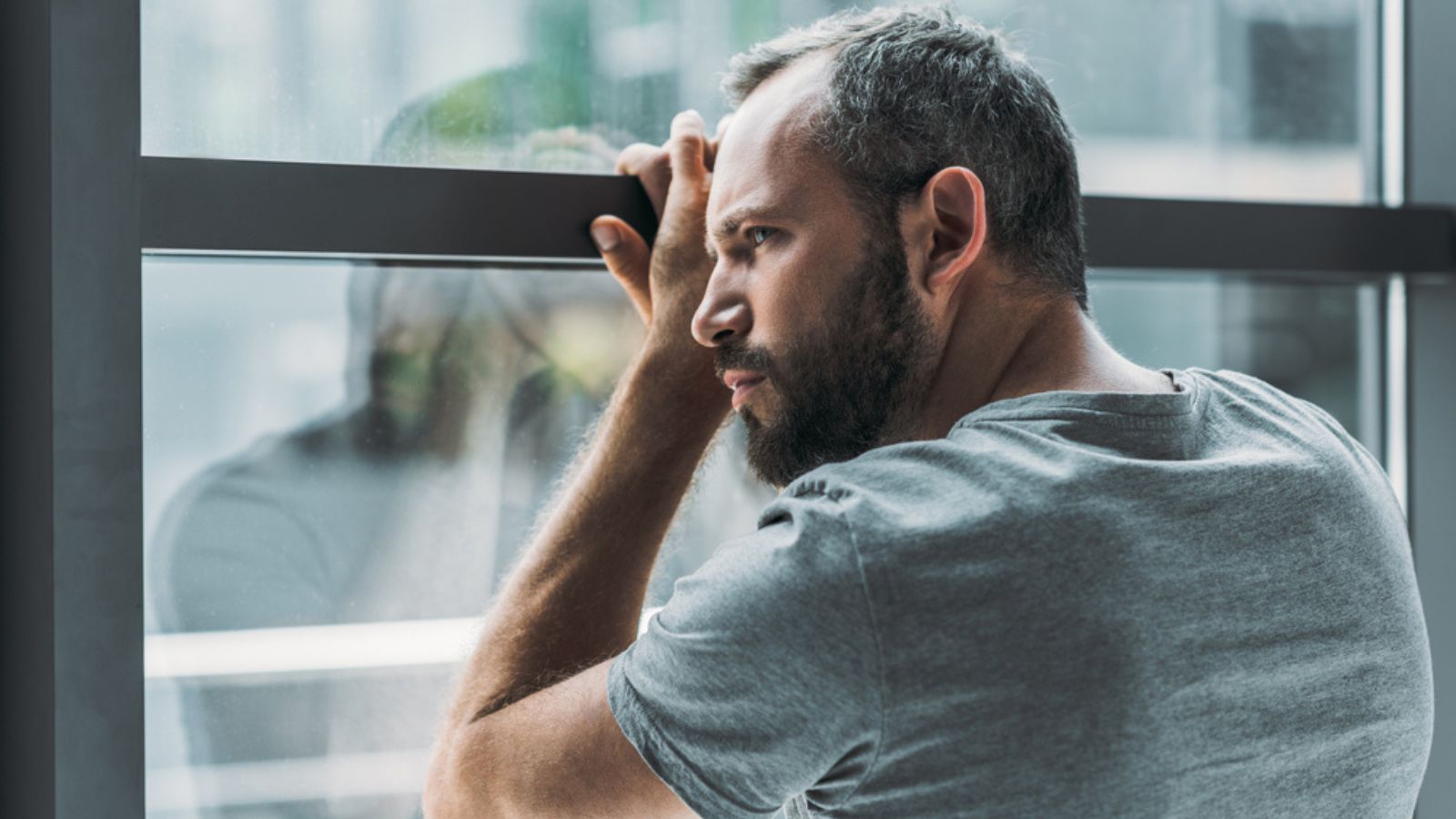 Upset bearded alone man leaning at window and looking through it