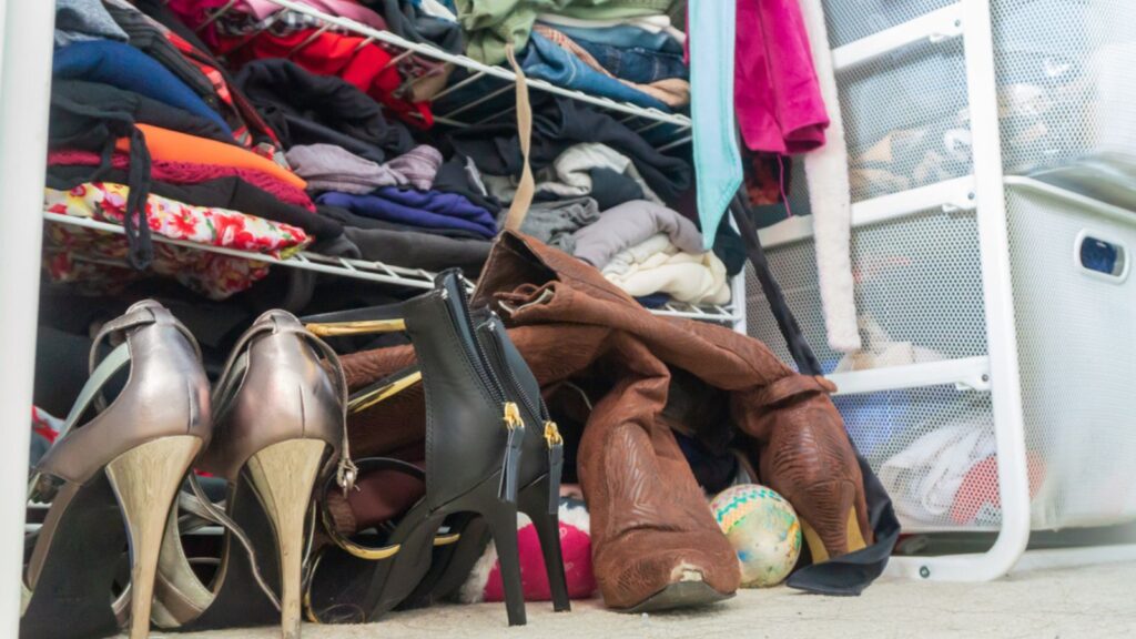Woman's closet with high heel shoes, stacked, folded clothes on shelves and part of robes hanging. Depicting closet organization, time to donate clothes, fashion lifestyle, consumerism.
