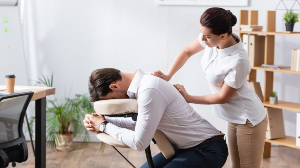 Side view of masseuse massaging back of businessman sitting on massage chair in office
