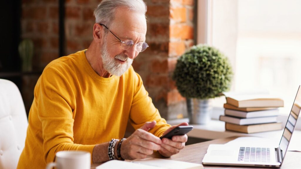 Cheerful aged freelancer smiling while sitting at a workplace with a phone in her hands in a home office working remote