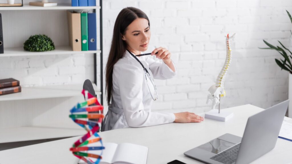 Doctor pointing at shoulder near spinal model during video call on laptop in clinic