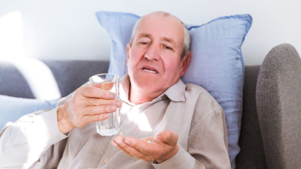Portrait of the old sick man lying on a bed and drinking pills at home