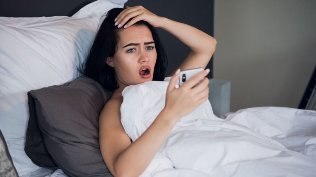Shocked young woman looking at mobile phone in bed at home