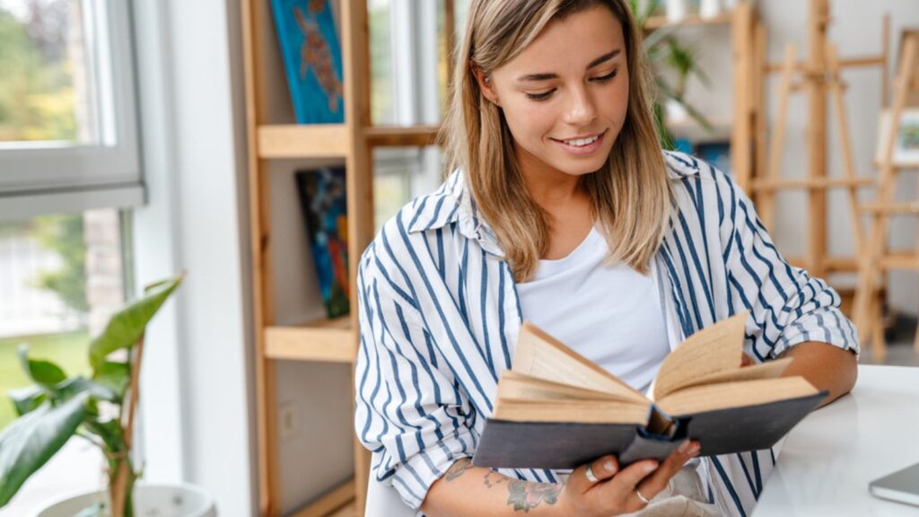 Smiling attractive young woman designer reading book while sitting at the workshop
