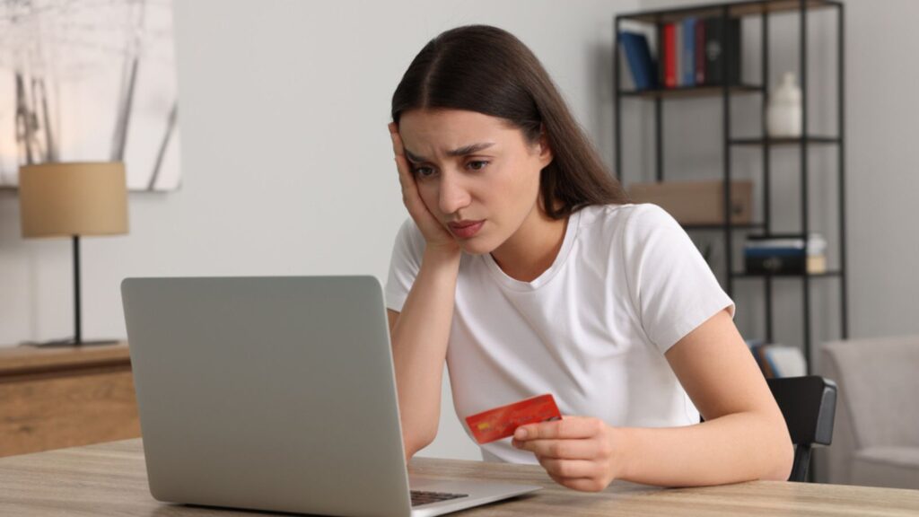 Upset woman with credit card using laptop at table indoors