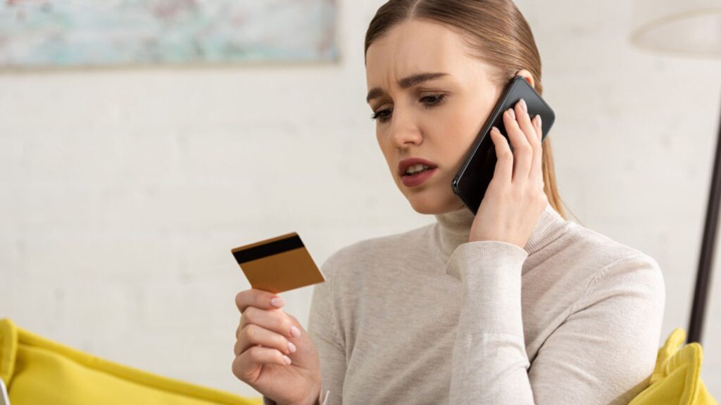Worried young woman holding credit card and talking on smartphone at home