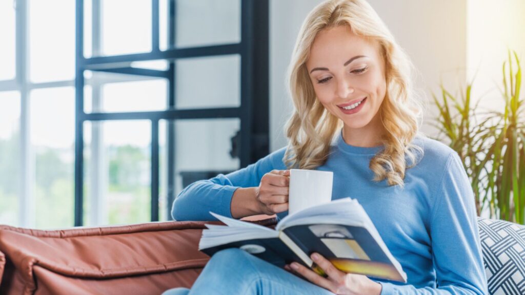 Young woman at home sitting on modern sofa relaxing in living room while reading a book and drinking coffee