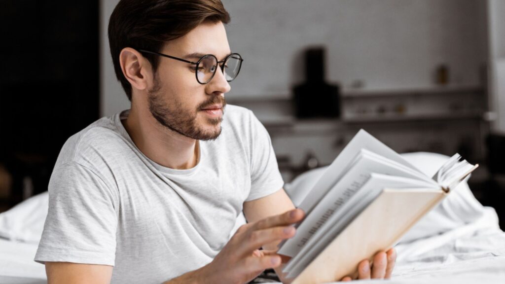 man in eyeglasses lying in bed and reading book in the morning