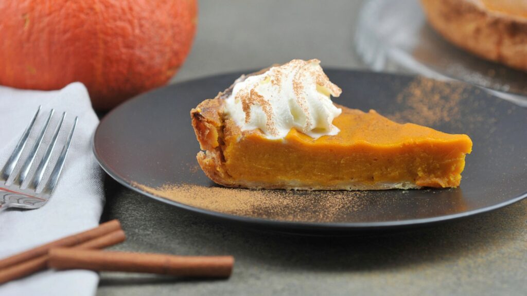 A piece of pumpkin pie on a black plate. The concept of a festive treat of the autumn harvest. Close-up. In the frame, cinnamon sticks, pumpkin, pie.