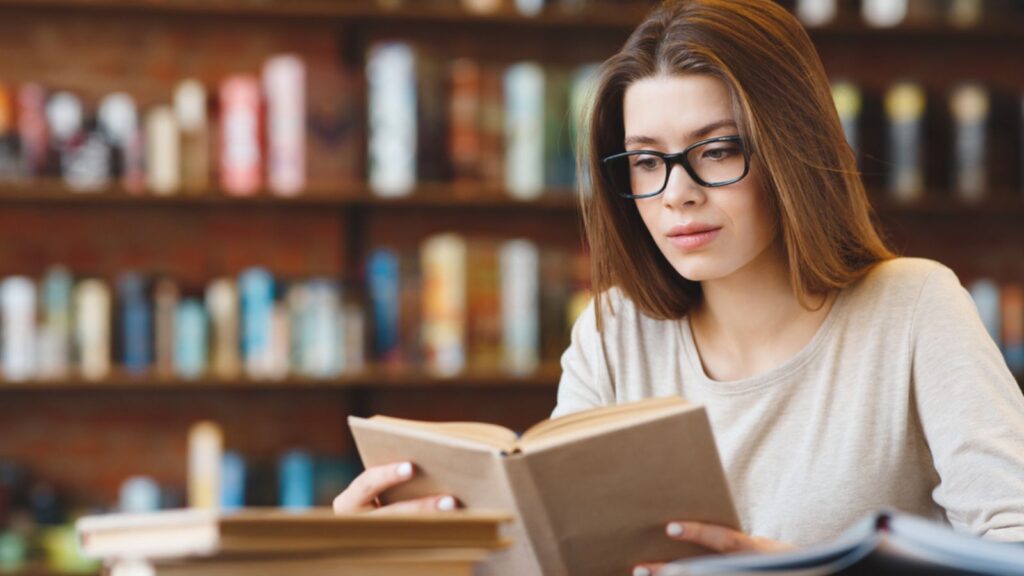 Attractive young woman in eyeglasses enjoying free time and reading a book