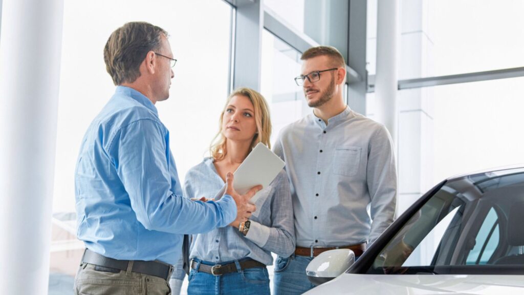 Car dealer helping young couple choose a new car by showing the advantages of the vehicle to the customer