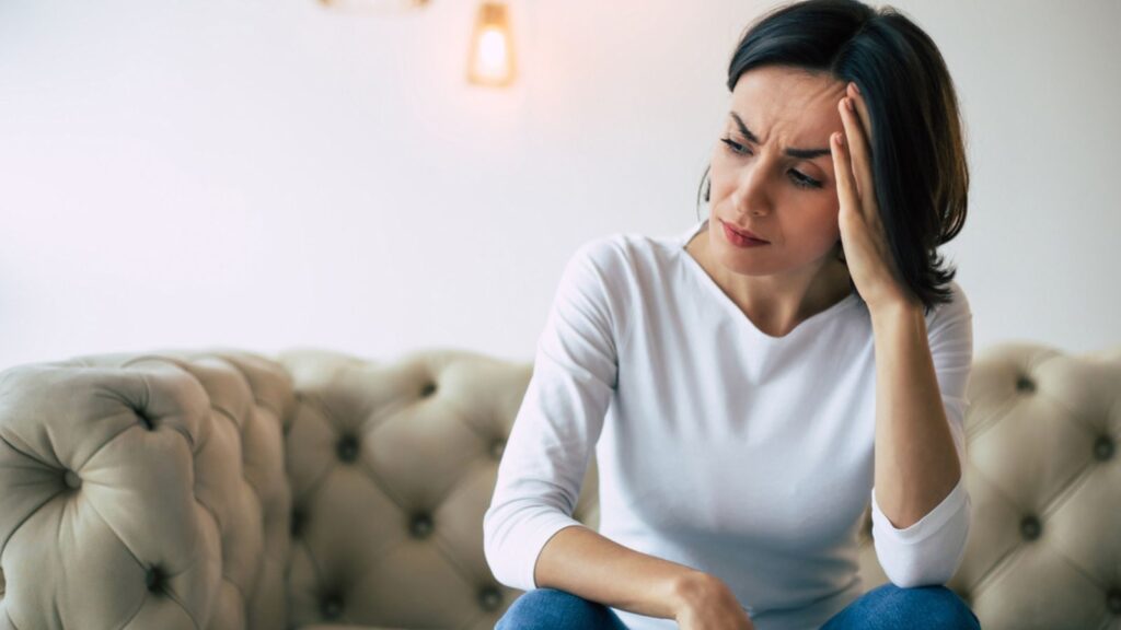 Close-up photo of a sad thinking woman who is sitting on a couch and touching her head while suffering from headache
