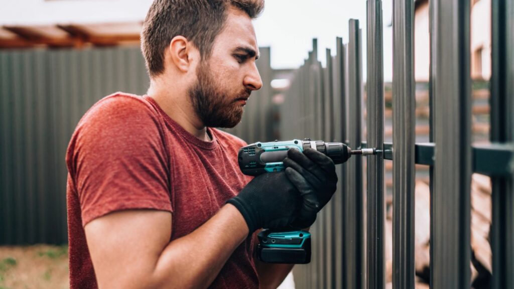 Construction worker using electrical screwdriver and mounting metal elements on fence