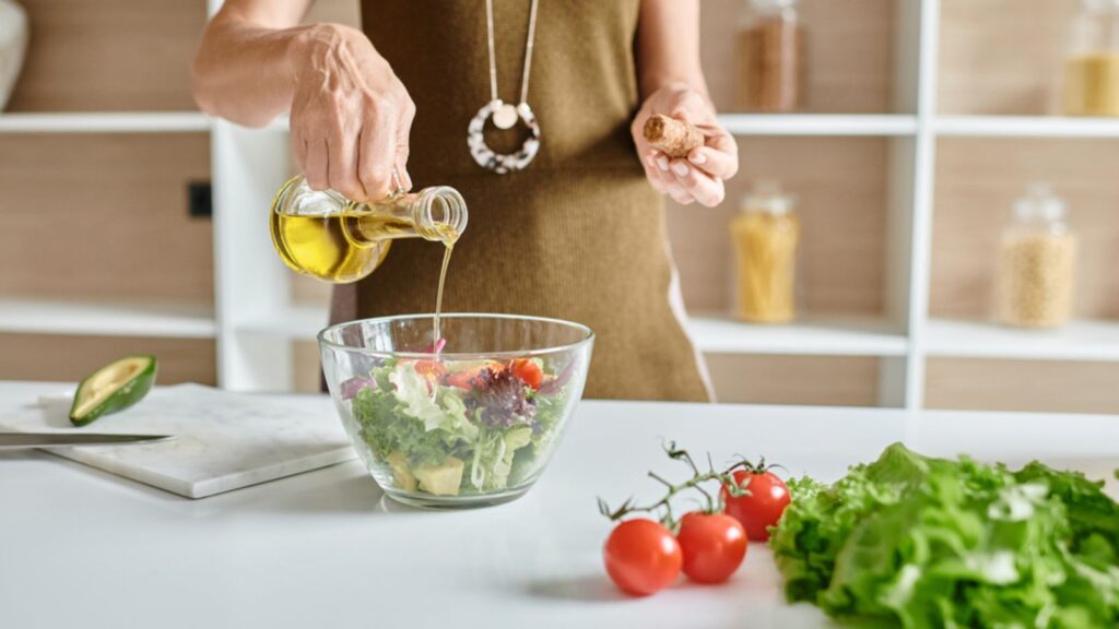 Cropped shot of woman pouring olive oil into glass bowl with salad near vegetables on countertop