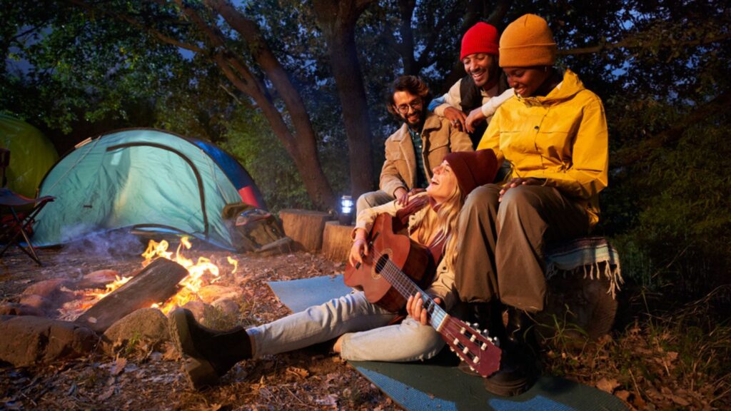 Group of friends with guitar near bonfire and camping tent outdoors overnight