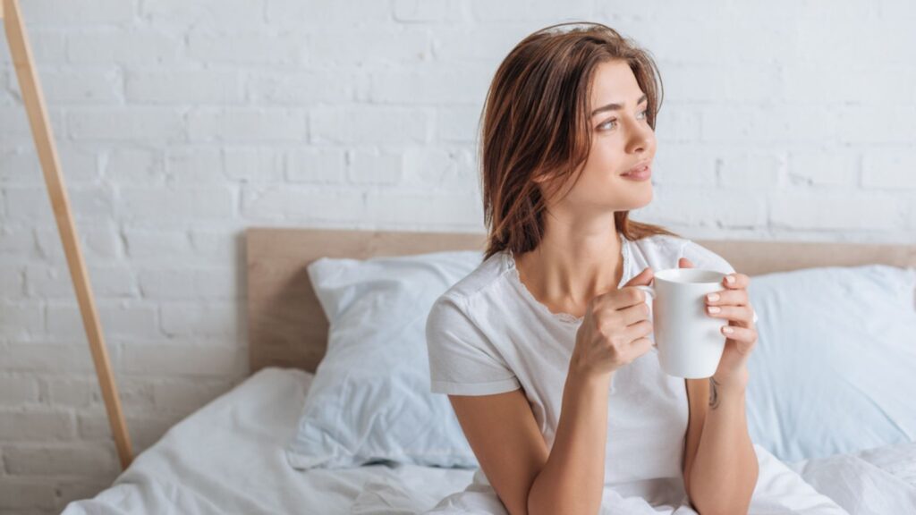 Happy young woman holding cup with tea in bedroom