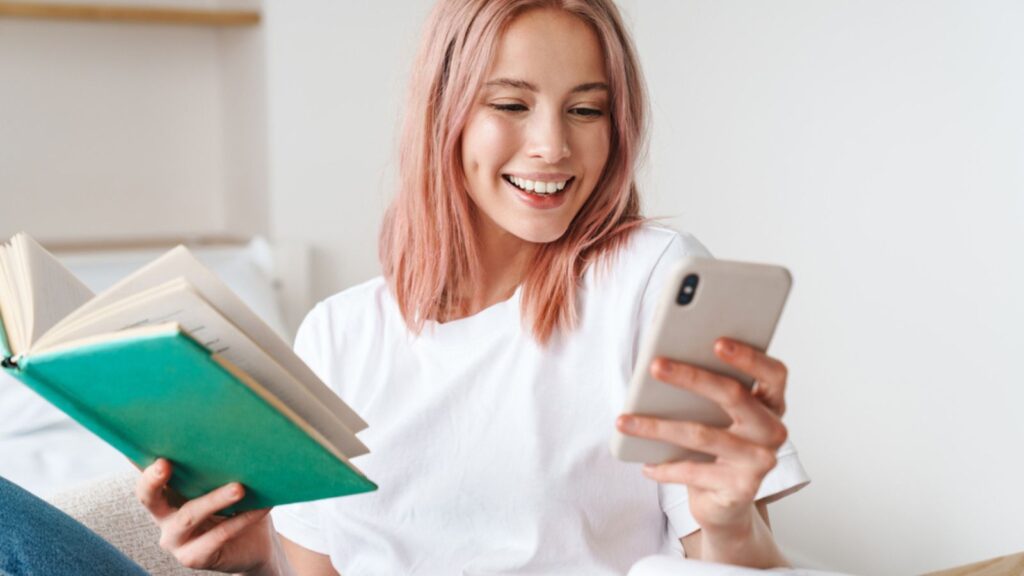 Image of joyful pretty woman with pink hair reading book and using cellphone while resting at home