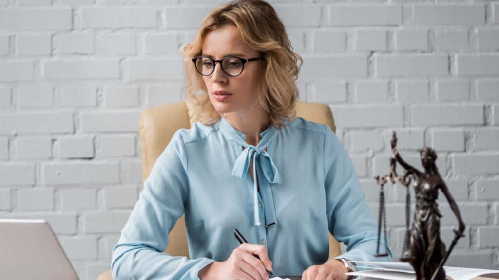 Serious woman lawyer in eyeglasses working with papers and using laptop at workplace