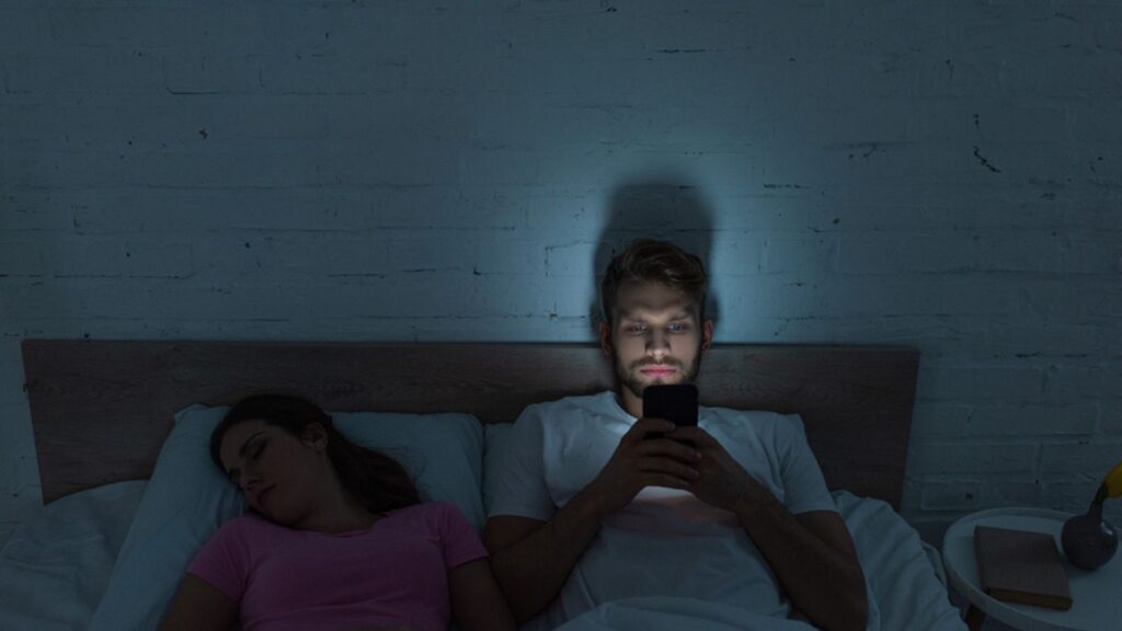 Smartphone depended man sitting on bed near sleeping girlfriend