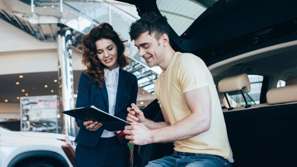 Smiling customer man and car dealer holding clipboard while standing near new car