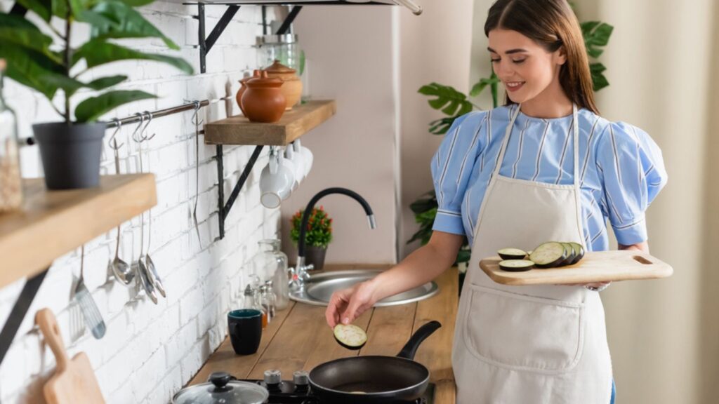 Young adult woman putting slices of eggplant in frying pan in kitchen