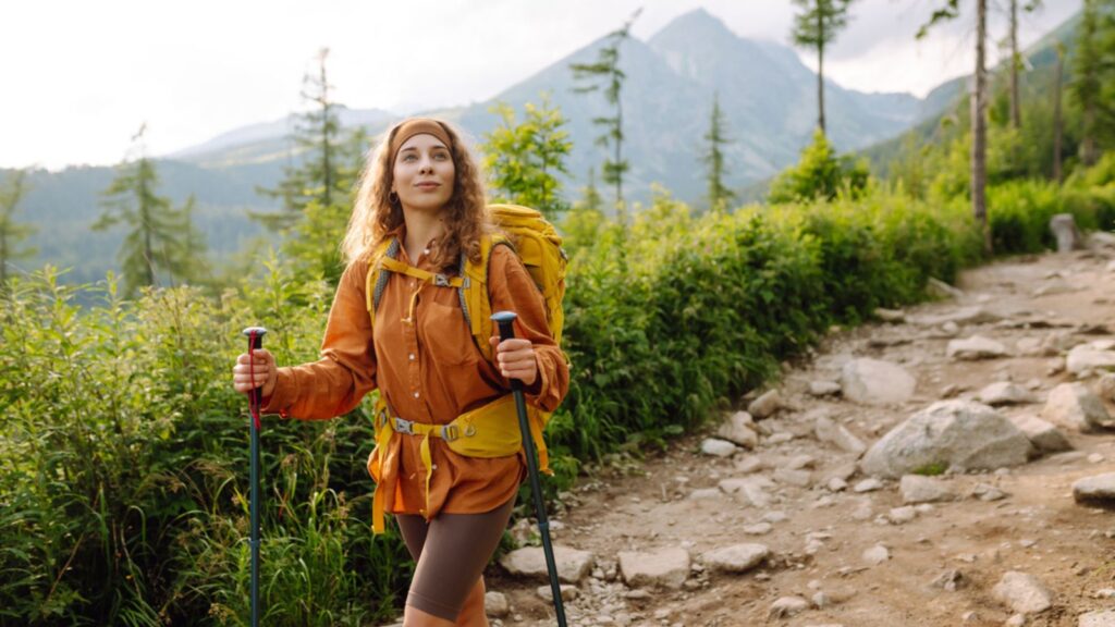 Young woman traveler with a yellow hiking backpack and hiking poles on trail among the mountains