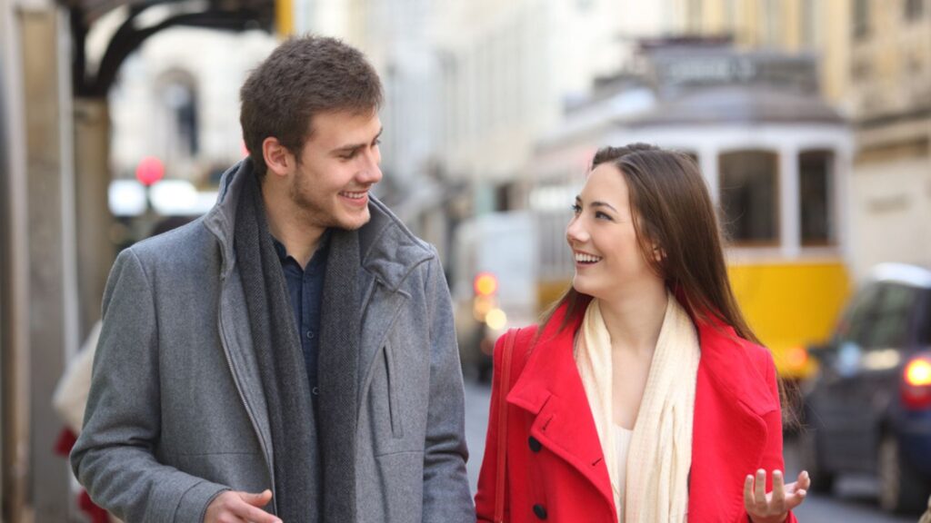 happy young couple in scarf and winter jacket walking in city talking laughing