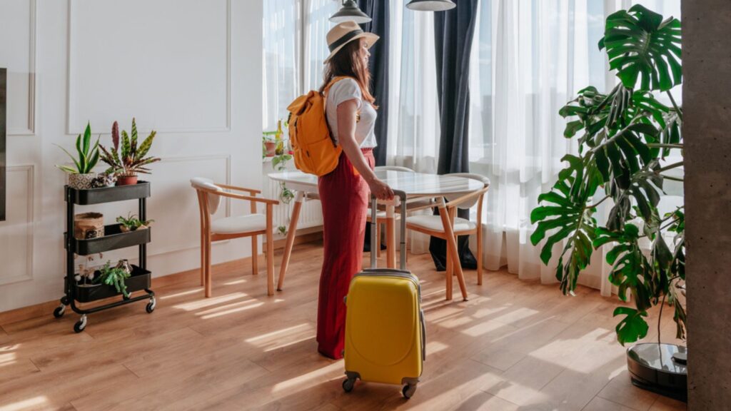 young traveler tourist woman looking out apartment window abroad with suitcase and backpack