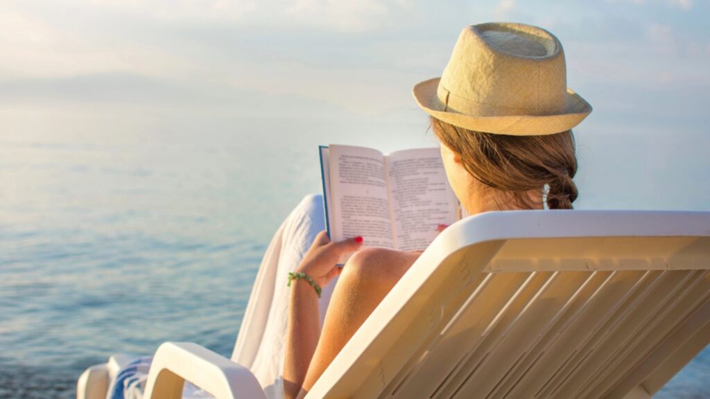 young woman sitting in beach chair by ocean reading a book at sunset