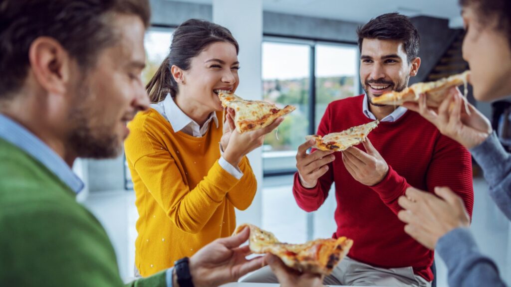 An excited multicultural group of businesspeople standing in the office eating pizza for lunch. Team building during office break. The smiling woman had just taken a bite of the pizza