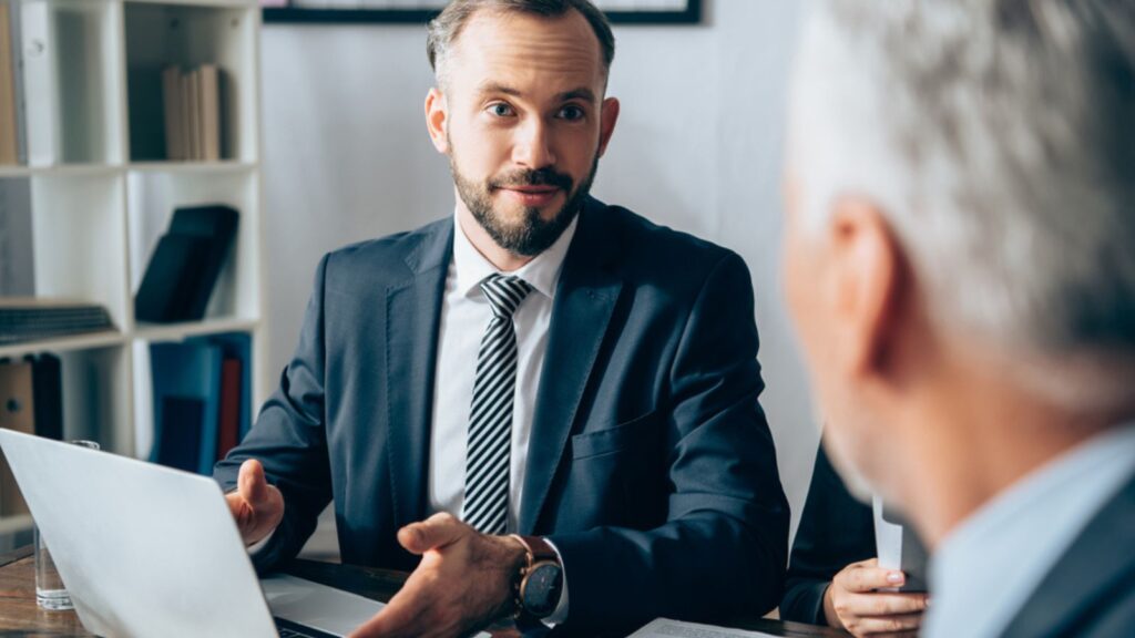 Businessman pointing at laptop near papers and investor on blurred foreground in office