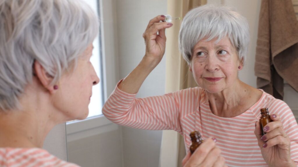 Close-up portrait of mature woman applying skincare oil in front of mirror in bathroom