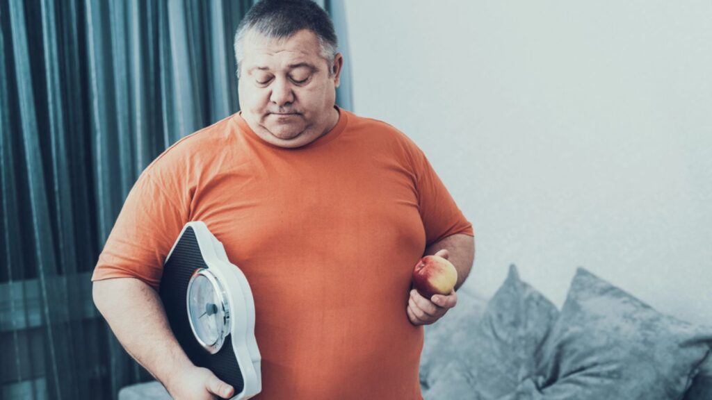 Fat Man in T-shirt with Weighter and Apple in Hands. Man with Bulimia. Unhealthy Lifestyle Concept. Sitting Man. Sad Man. Treatment of Overweight. Man with Overweight.
