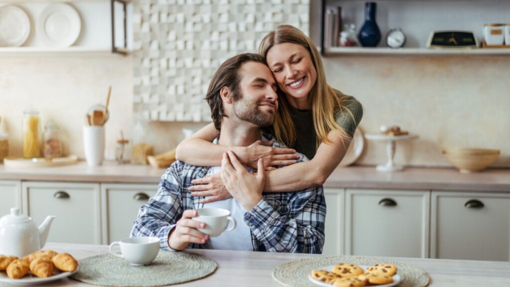 Glad young european lady hugging male with stubble drink coffee, enjoy free time in modern kitchen interior