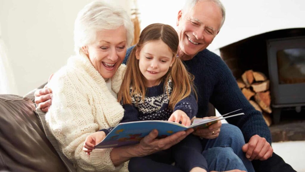 Grandparents Sitting On Sofa With Granddaughter At Home Reading Book