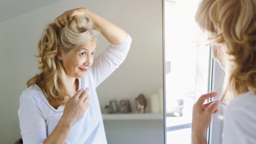 Happy beautiful senior woman cheking her hair in front of mirror at home