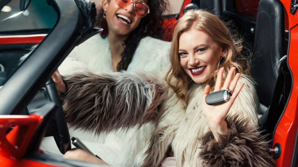 Happy stylish multiethnic women in fur coats holding car keys while sitting in luxury red car
