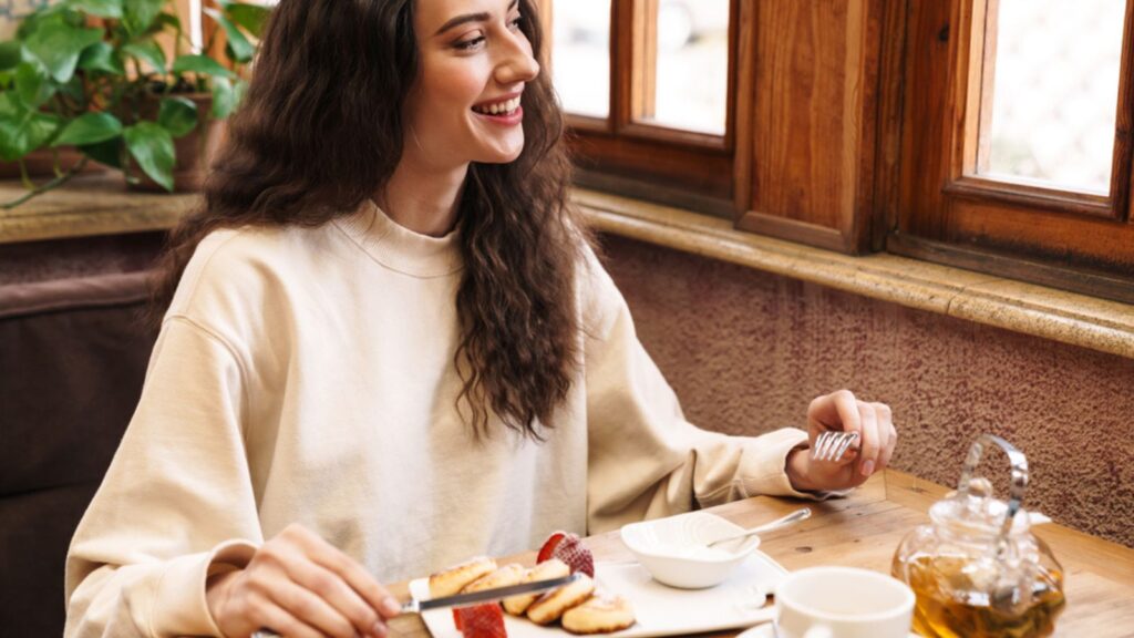 Image of happy beautiful young woman smiling and eating pancakes while sitting in cozy cafe indoors