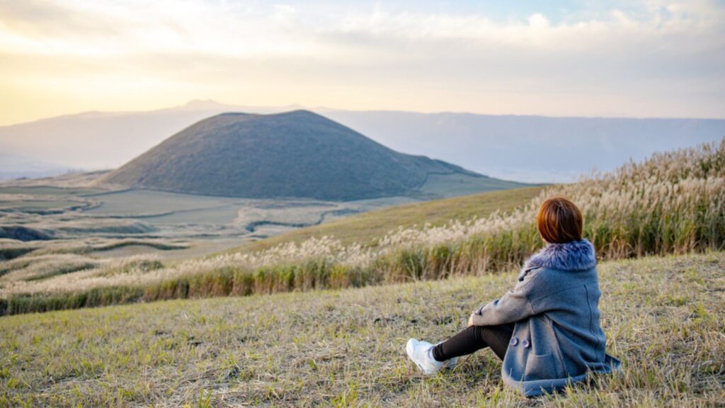 Lovely cute japanese girl sitting next to Aso active volcano background with smoke at Mount Aso Nakadake, Kumamoto, kyushu, Japan