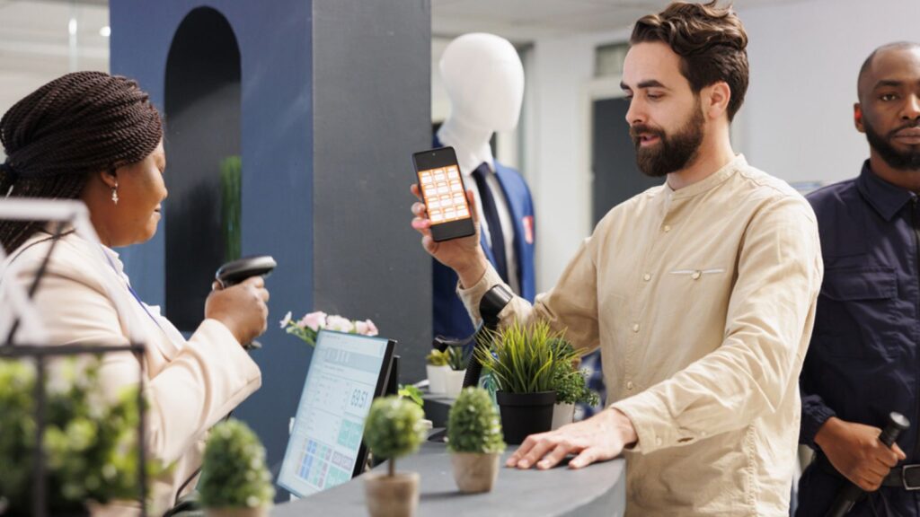 Man customer holding smartphone showing mobile discount code to saleswoman while shopping in clothing store. Cashier scanning promo coupon while serving shopper at checkout counter