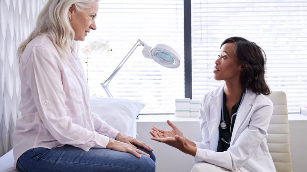 Mature Woman In Consultation With Female Doctor Sitting On Examination Couch In Office